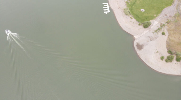 A drone image of Island Park Reservoir. Two thirds of the image is gray colored water, with a boat moving toward the top left corner. The right third of the photo shows a dock and dry land with a small lawn.