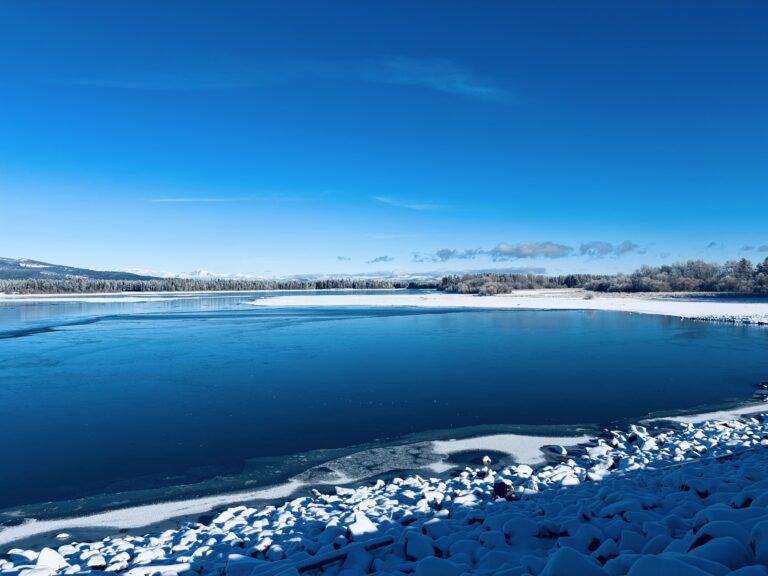 A reservoir with calm blue water. There is snow along the shores. The color of the sky matches the blue of the lake and is free of clouds. Trees sit in the distance.