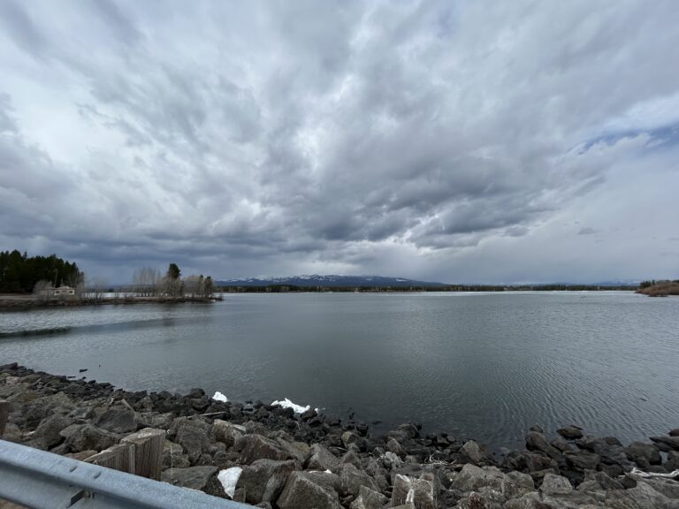 A photo taken of Island Park Reservoir while standing on the dam and looking north. The sky is cloudy, Mt Sawtelle sits with little snow in the background, and the reservoir is open water (save for a few small sheets of ice in the distance).
