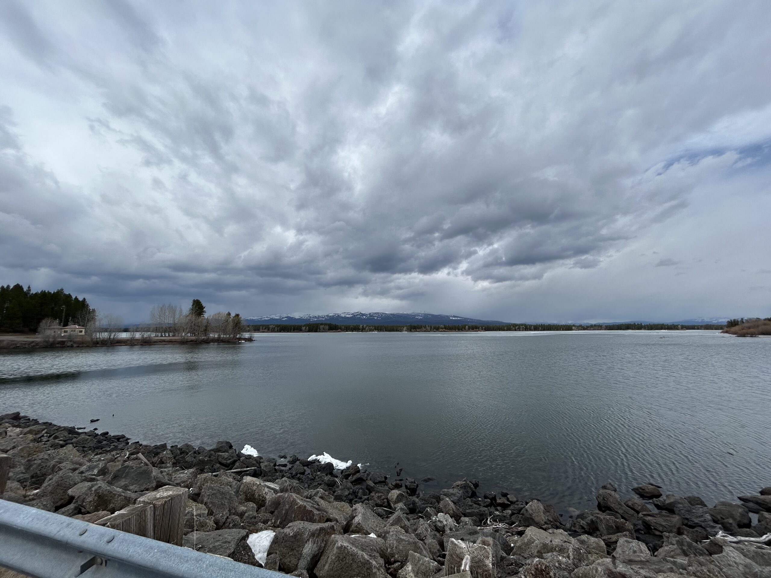 A photo taken of Island Park Reservoir while standing on the dam and looking north. The sky is cloudy, Mt Sawtelle sits with little snow in the background, and the reservoir is open water (save for a few small sheets of ice in the distance).