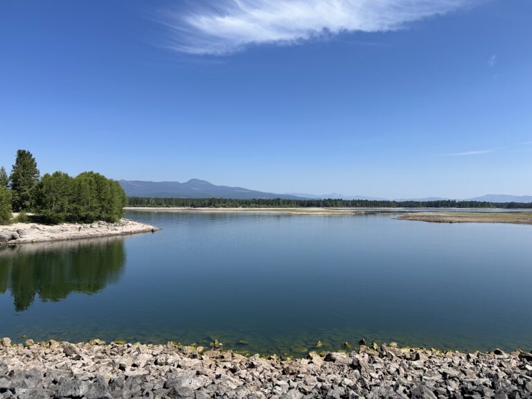 The blue water of Island Park Reservoir under a blue sky with a single white cloud. Sawtelle sits in the background and you can see rocky edges of the reservoir as the water levels have dropped.