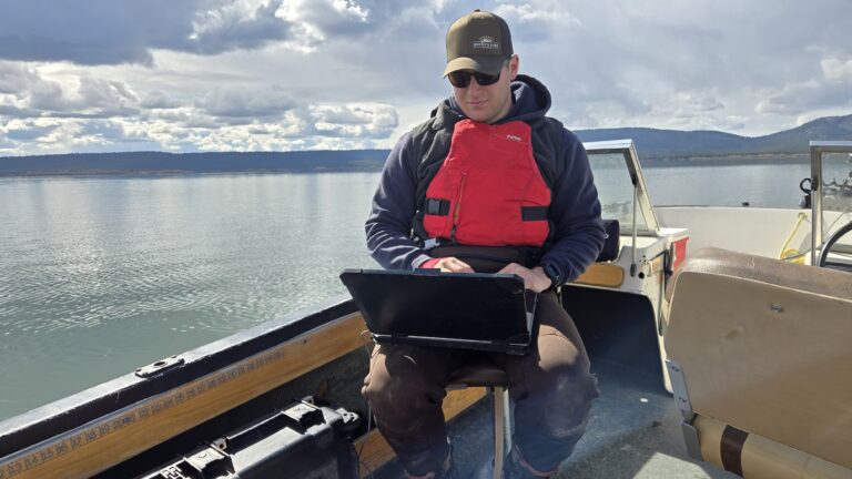 A person wearing a red life jacket, sunglasses, and brimmed hat sits on a boat with a laptop. A lake and mountain are in the background.