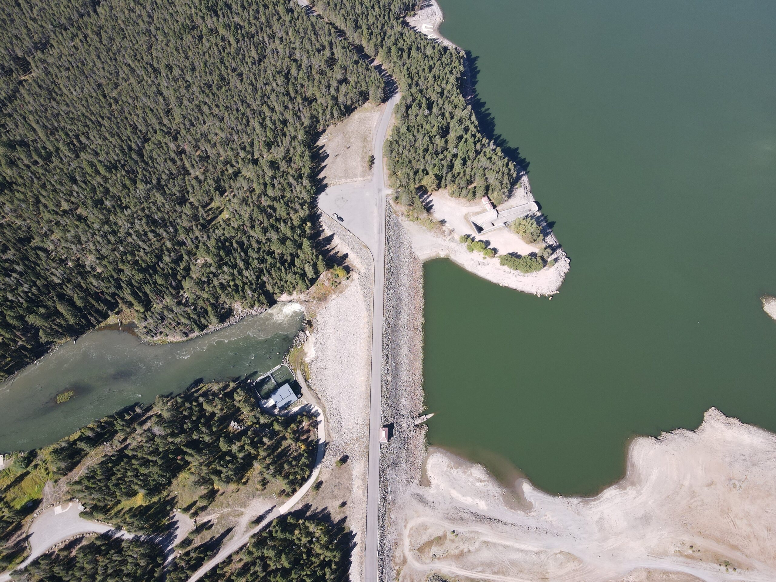 Drone image of Island Park Dam, where a low lake level is visible on the right (above the dam) and turbid outflow is visible on the left (below the dam).