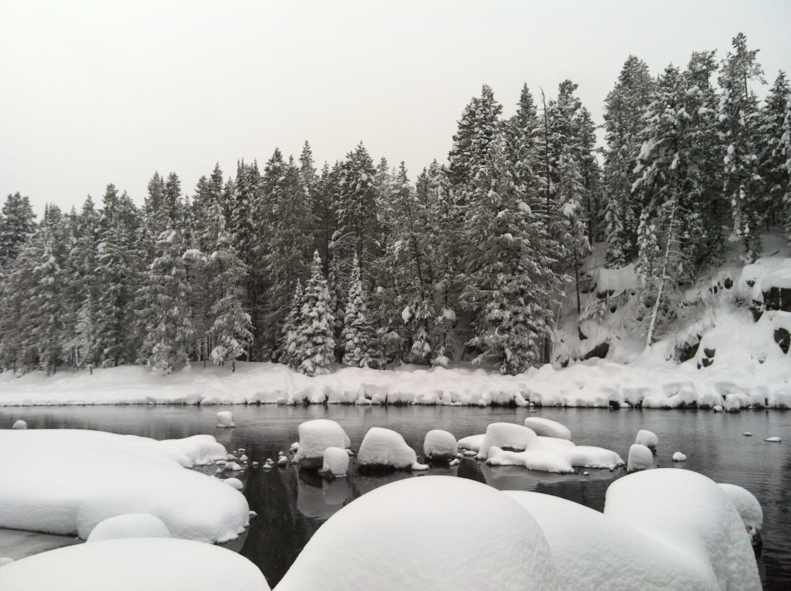 A river surrounded by snow. There are rocks in the foreground pillowy with snow. The river flows from right to left in the middle. On the other side sits needled trees and a snow covered bank. The photo almost appears to be black and white.